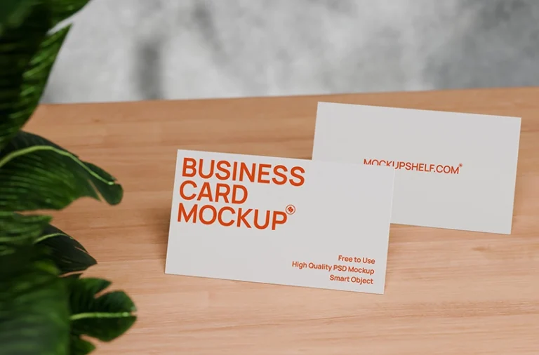 Free business card mockup on desk PSD with two cards on a wooden table and green plant leaves